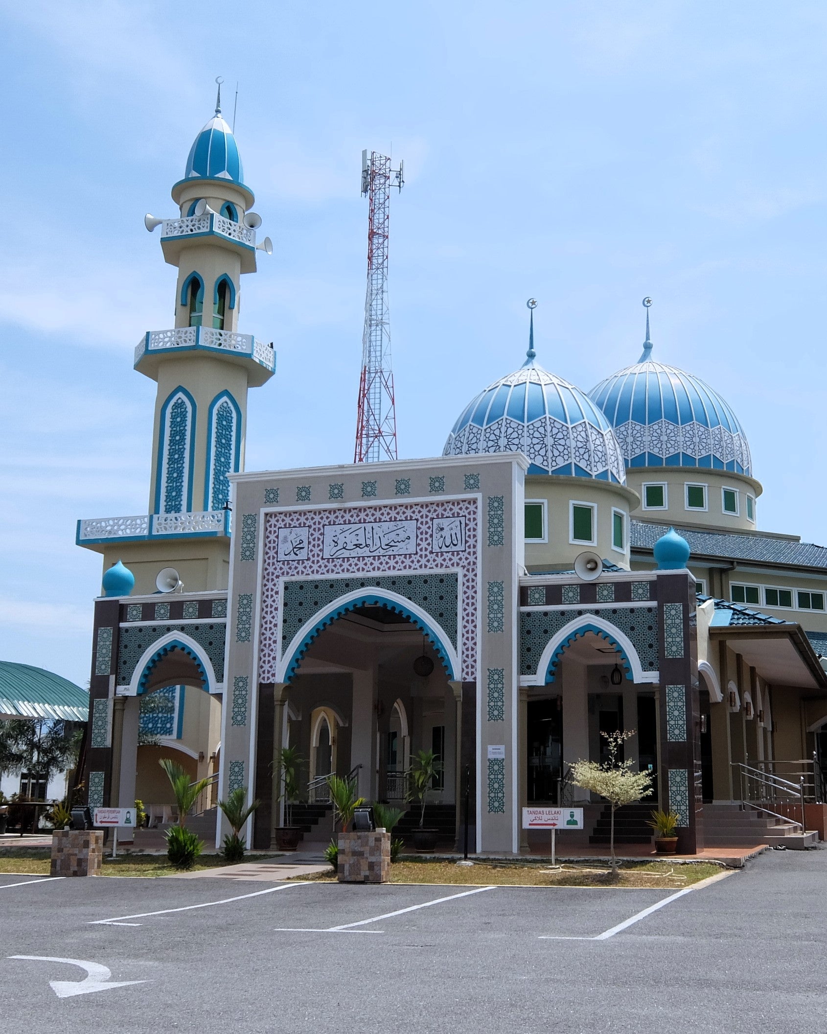 A large mosque with blue and white domes and minarets, photographed outdoors under a pale blue sky. The building has ornate arches, decorative tilework, and a paved parking area in front.