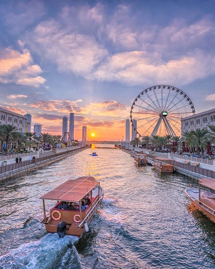 A scenic sunset view of a canal with boats, a large Ferris wheel, modern buildings, and palm trees under a dramatic cloudy sky.