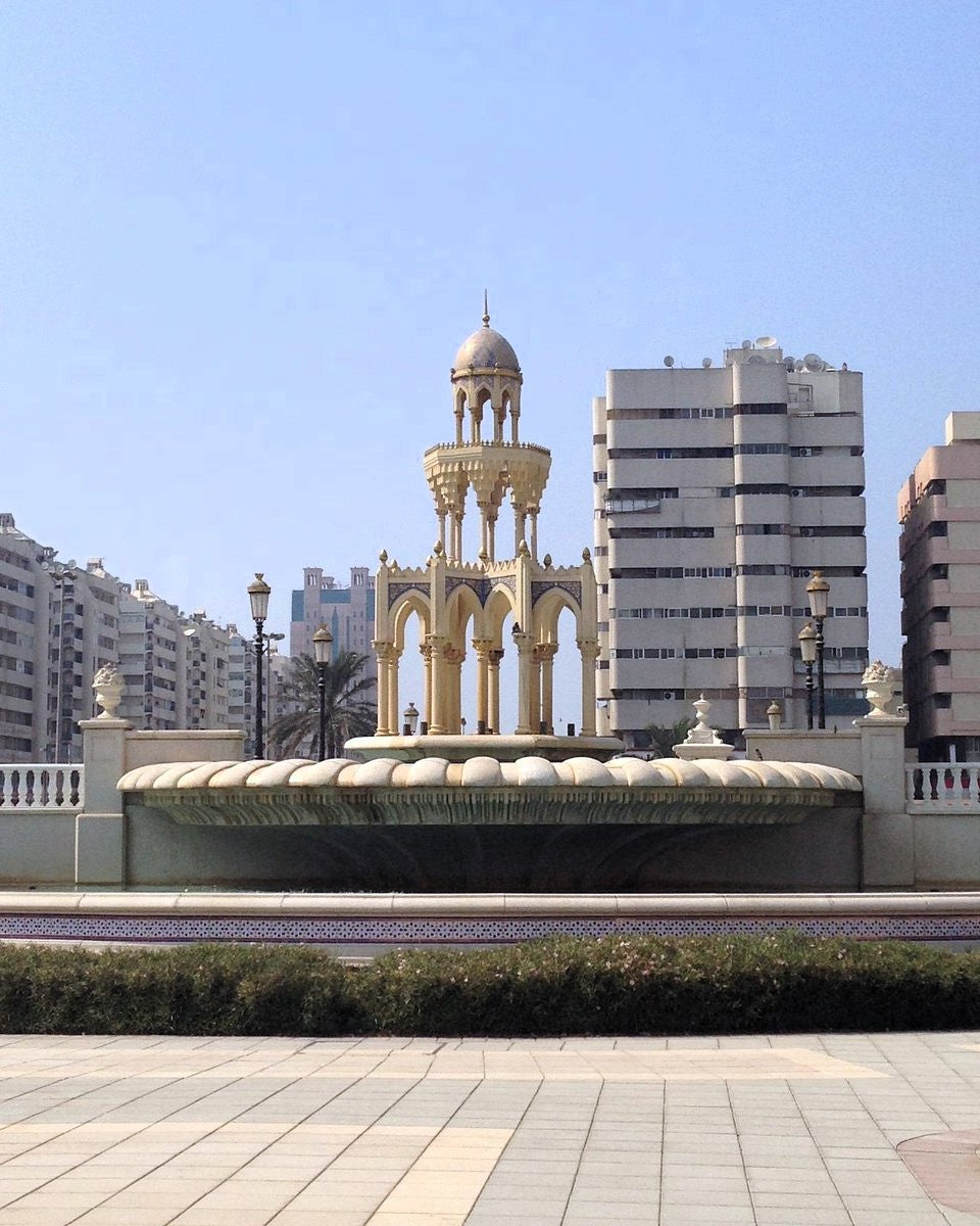 A golden ornamental fountain structure with a domed top and arched columns, set in an urban plaza with buildings and a paved walkway in the foreground.