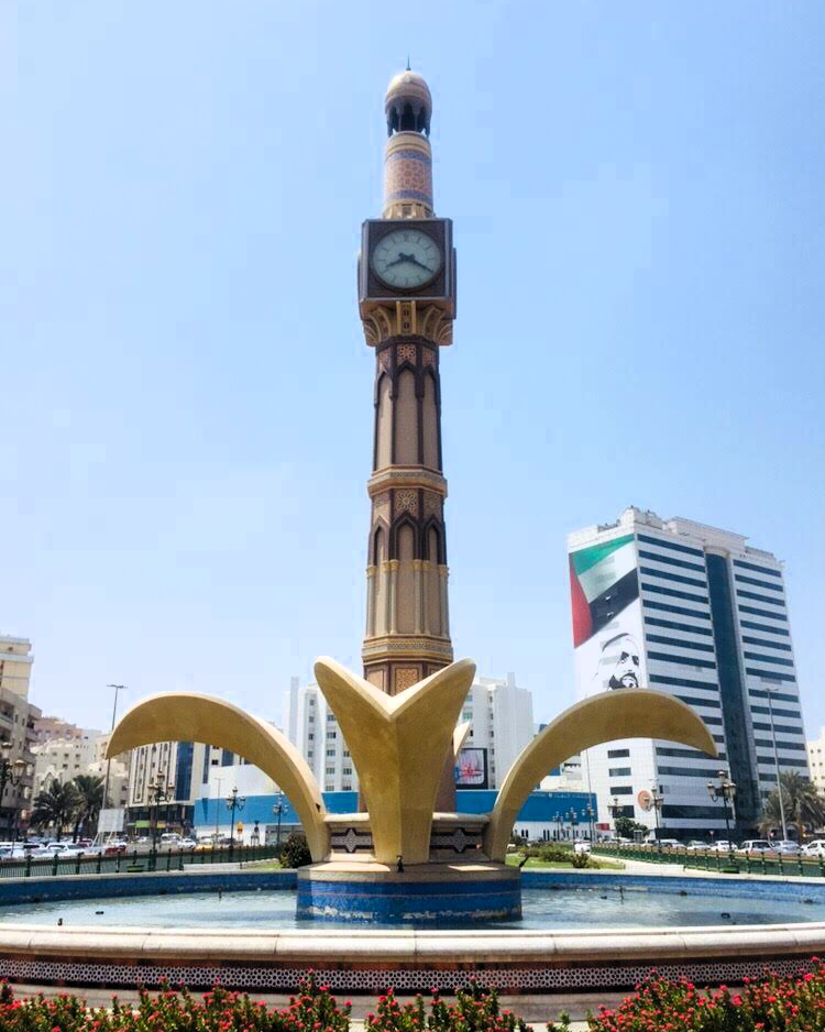 A tall clock tower monument in an urban plaza, with a fountain base featuring curved golden arches and surrounding buildings under a clear blue sky.