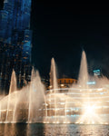 A nighttime city scene with a large illuminated fountain display in the foreground and a tall blue-lit skyscraper on the left. The fountain jets shoot upward into a dark sky, with bright lights reflecting on the water surface.