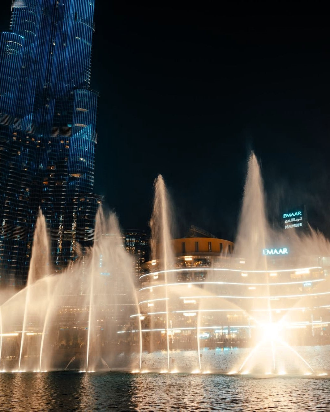A nighttime city scene with a large illuminated fountain display in the foreground and a tall blue-lit skyscraper on the left. The fountain jets shoot upward into a dark sky, with bright lights reflecting on the water surface.