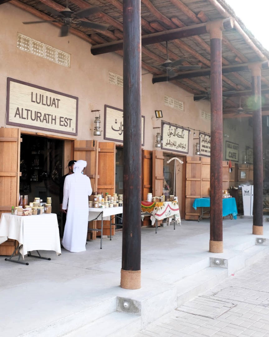 A traditional Middle Eastern storefront with wooden pillars, Arabic and English signage reading 'LULUAT ALTURATH EST', and a person in a white robe standing near tables displaying goods on a covered porch.
