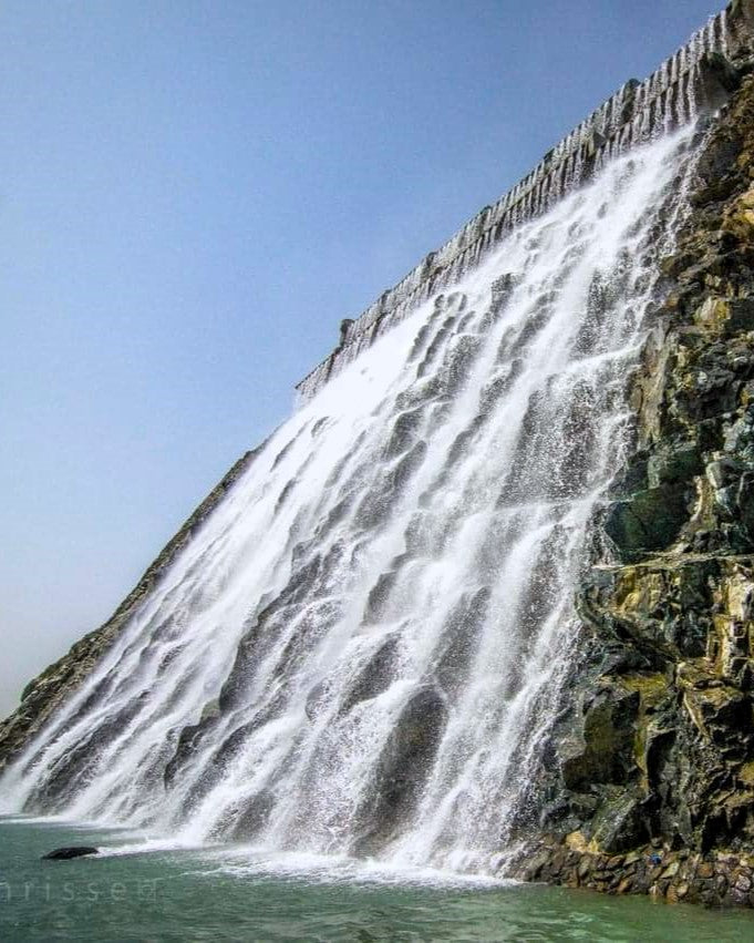A large waterfall cascades down a steep rocky cliff into turquoise water under a clear blue sky.