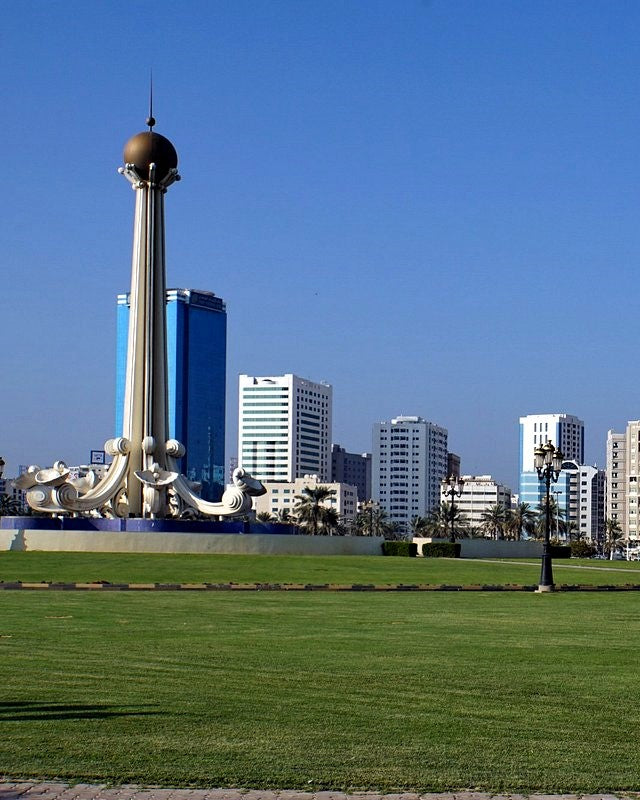 A scenic view of a city skyline with a prominent white monument featuring a gold sphere on a tall pole, situated on a large green lawn under a clear blue sky.