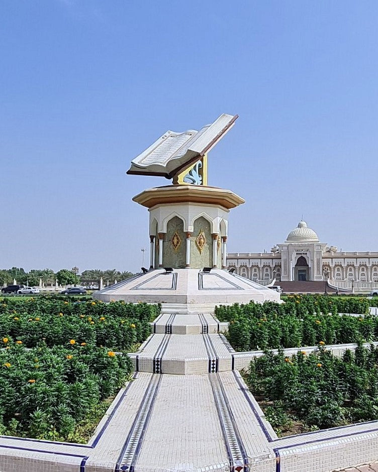 A large outdoor architectural monument featuring an open book sculpture mounted above a domed pavilion, with a paved walkway leading up the center. The scene is surrounded by manicured green hedges and a pale blue sky, with a domed building visible in the background.