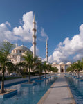 A scenic photograph of a large white mosque with two tall minarets, surrounded by palm trees and a long reflecting pool under a bright blue sky with white clouds.