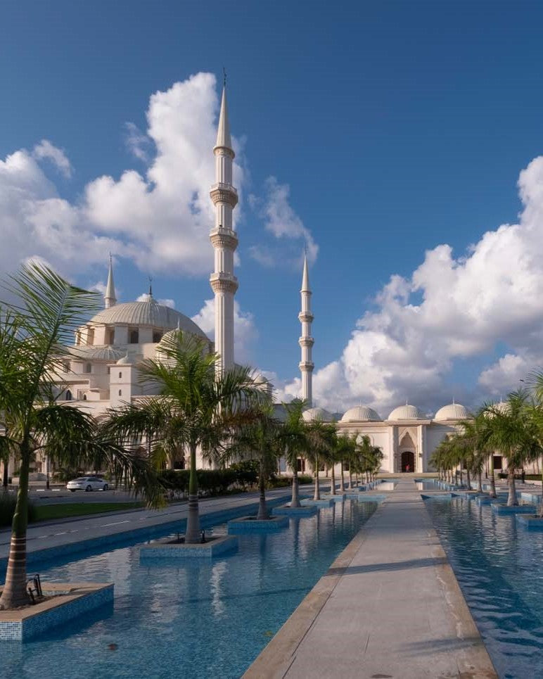A scenic photograph of a large white mosque with two tall minarets, surrounded by palm trees and a long reflecting pool under a bright blue sky with white clouds.