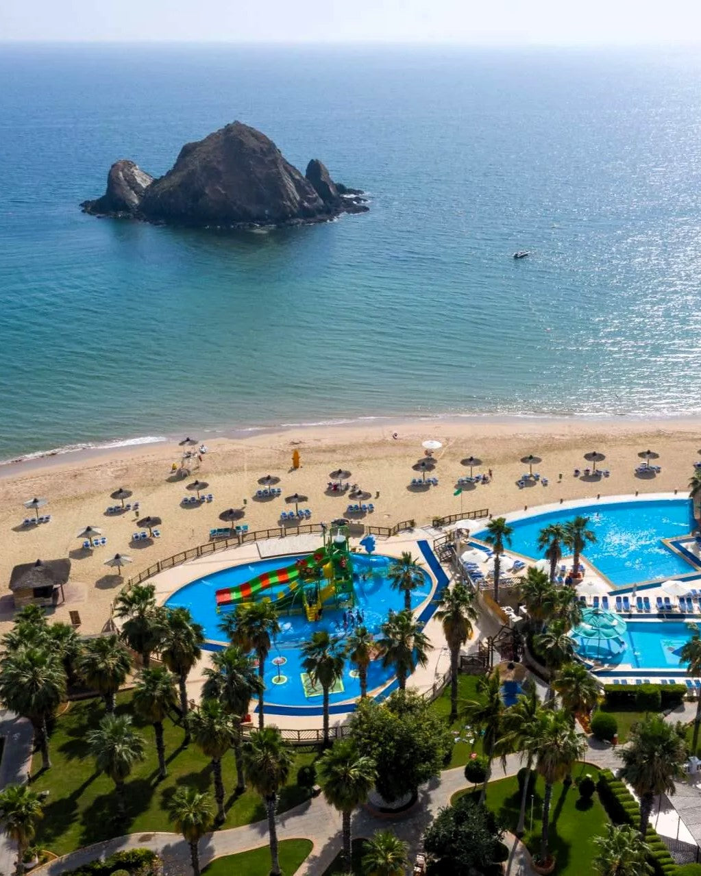 Aerial view of a coastal resort with a sandy beach, blue ocean, a rocky island in the water, and resort pools surrounded by palm trees and umbrellas.