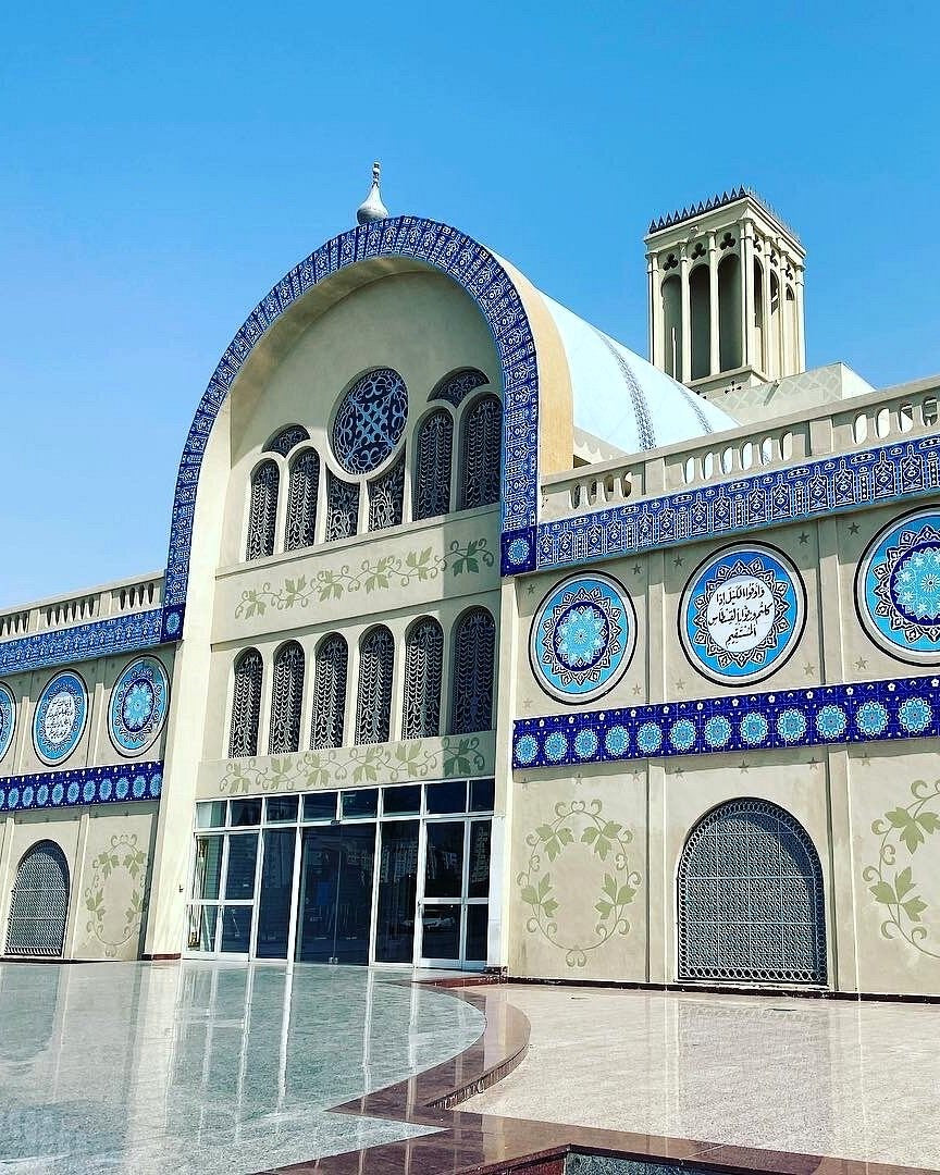 An ornate mosque facade with beige walls, blue mosaic tile detailing, arched windows, a central dome structure, and a clear blue sky above.