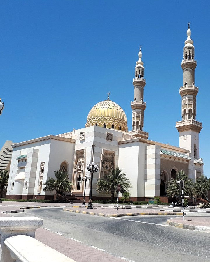 A large white mosque with a prominent gold dome and two tall minarets, photographed outdoors under a clear blue sky with paved streets and a few palm trees in the foreground.